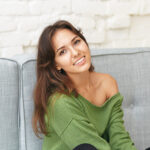 Indoor shot of attractive carefree young mixed race lady with brunette hair relaxing on comfortable couch in living room surfing websites and social networks using mobile phone, smiling happily